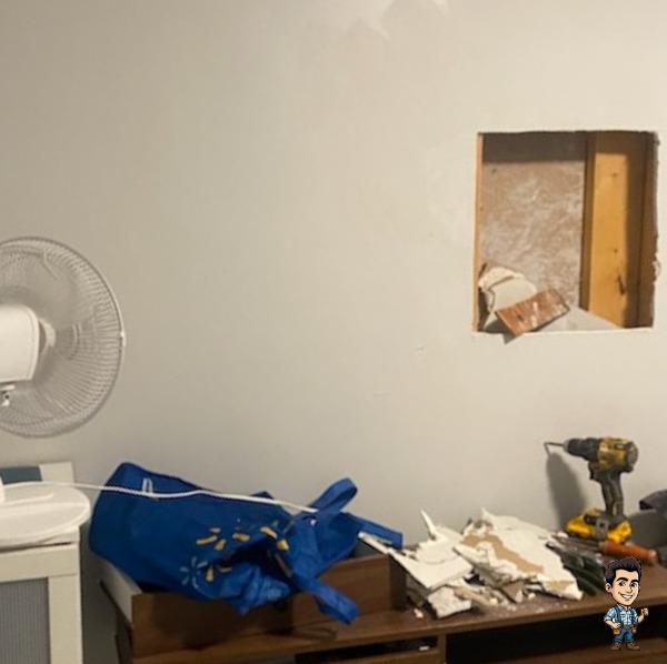 Bedroom wall with a large square hole cut out and drywall debris on a dresser before repair